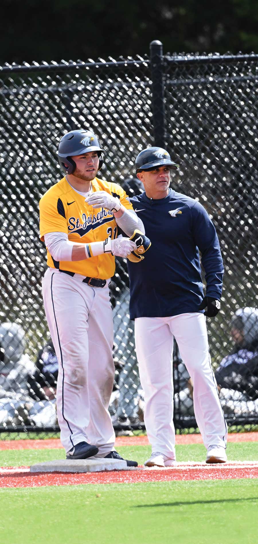 A portrait photograph of St. Joseph's Long Island Golden Eagles assistant baseball coach Andrew "Asch" Aschettino (pictured on the right) next to a Golden Eagles baseball player in their respective uniforms (navy blue and yellow) on first base as they glance over at the home plate area of the baseball field