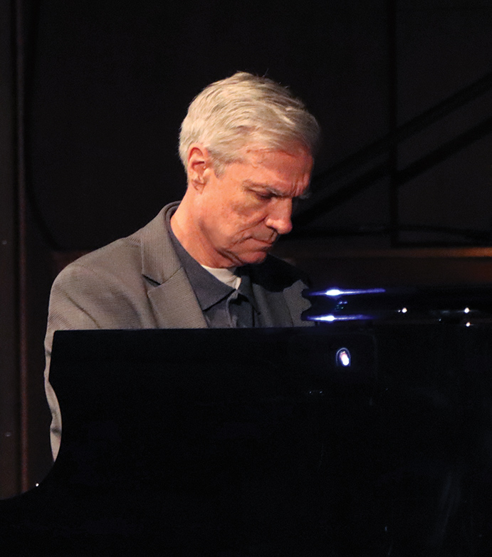 St. Joseph’s University, New York President Donald R. Boomgaarden (wearing a grey colored business suit jacket, dark navy blue button-up dress shirt, and glancing downward at the piano keys)