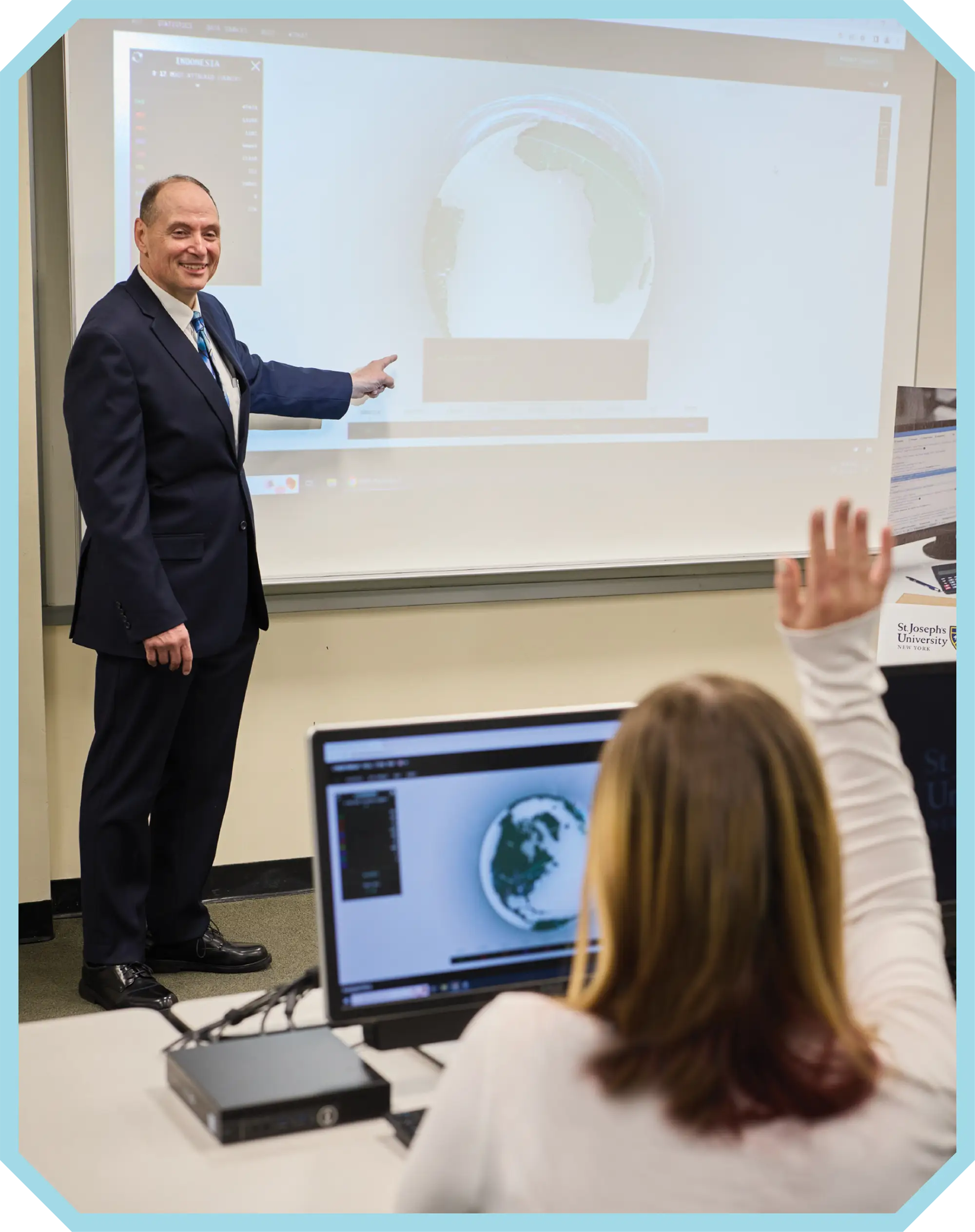 man in suit teaching a class at the projector screen while a student raises their hand