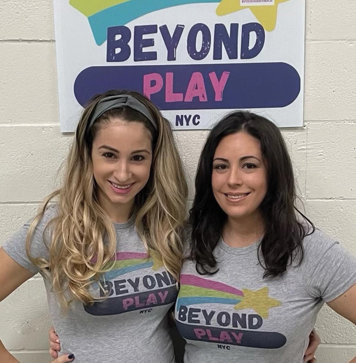 Madeline Romano ’09 and Mary Vitale ’10 stand together smiling and wearing "Beyond Play NYC"  while standing in front of a poster that reads "Beyond Play NYC"