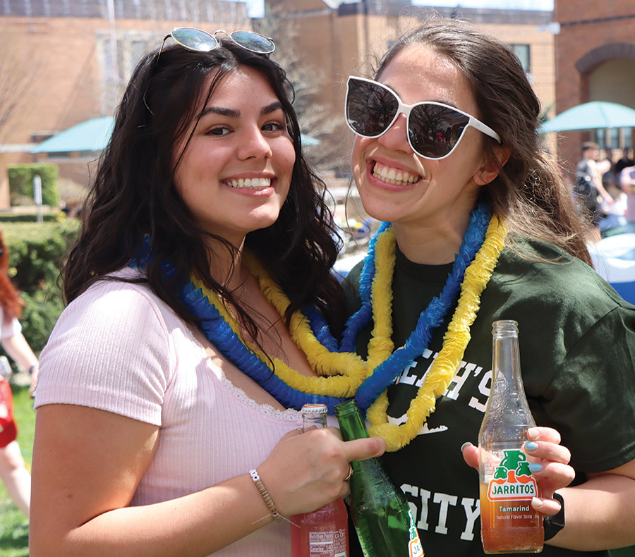 Two St. Joseph's University, New York students are smiling and posing for a picture together outside on campus at the Spring Fling Week event as one of the students is wearing sunglasses on top of their head, a blue/yellow Hawaiian style lei around her neck, and a pink t-shirt as she carries an orange flavored Jarritos fruit soda brand while the other student is wearing a dark forest green St. Joseph's University, New York t-shirt with a blue/yellow Hawaiian style lei around her neck and has white sunglasses on/covering her eyes while she carries a orange flavored Jarritos fruit soda brand bottle and wearing a watch on her arm