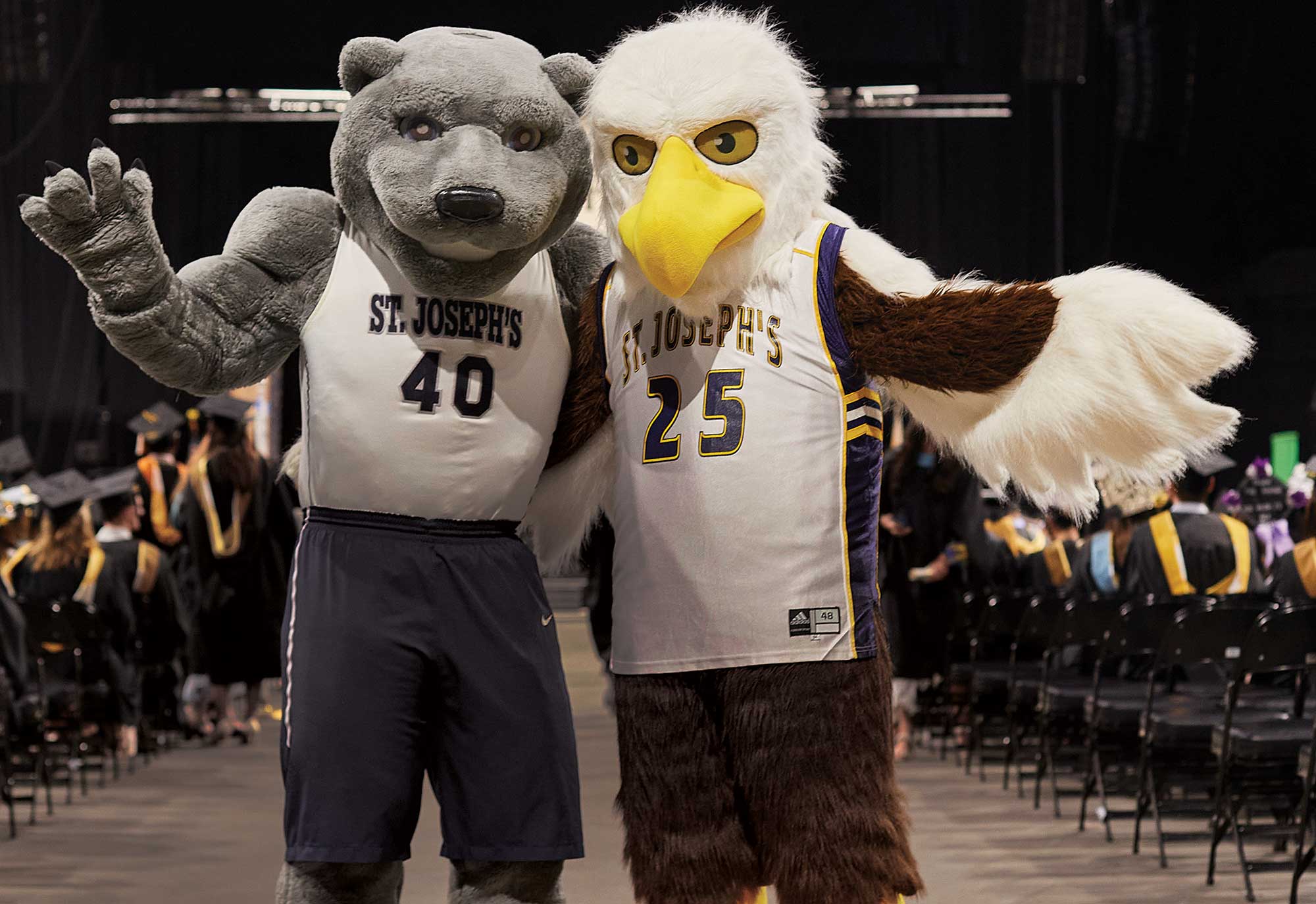 Landscape close-up photograph perspective of St. Joseph's University New York mascots Vandy (dressed on the left in a grey colored bear shaped costume suit with a white/grey/navy blue St. Joseph's basketball jersey and navy blue basketball shorts) and Hot Wyngz (dressed on the right in a brown/white colored eagle shaped costume suit with a white/yellow/navy blue St. Joseph's basketball jersey) posing for a picture next to each other at a graduation ceremony indoors