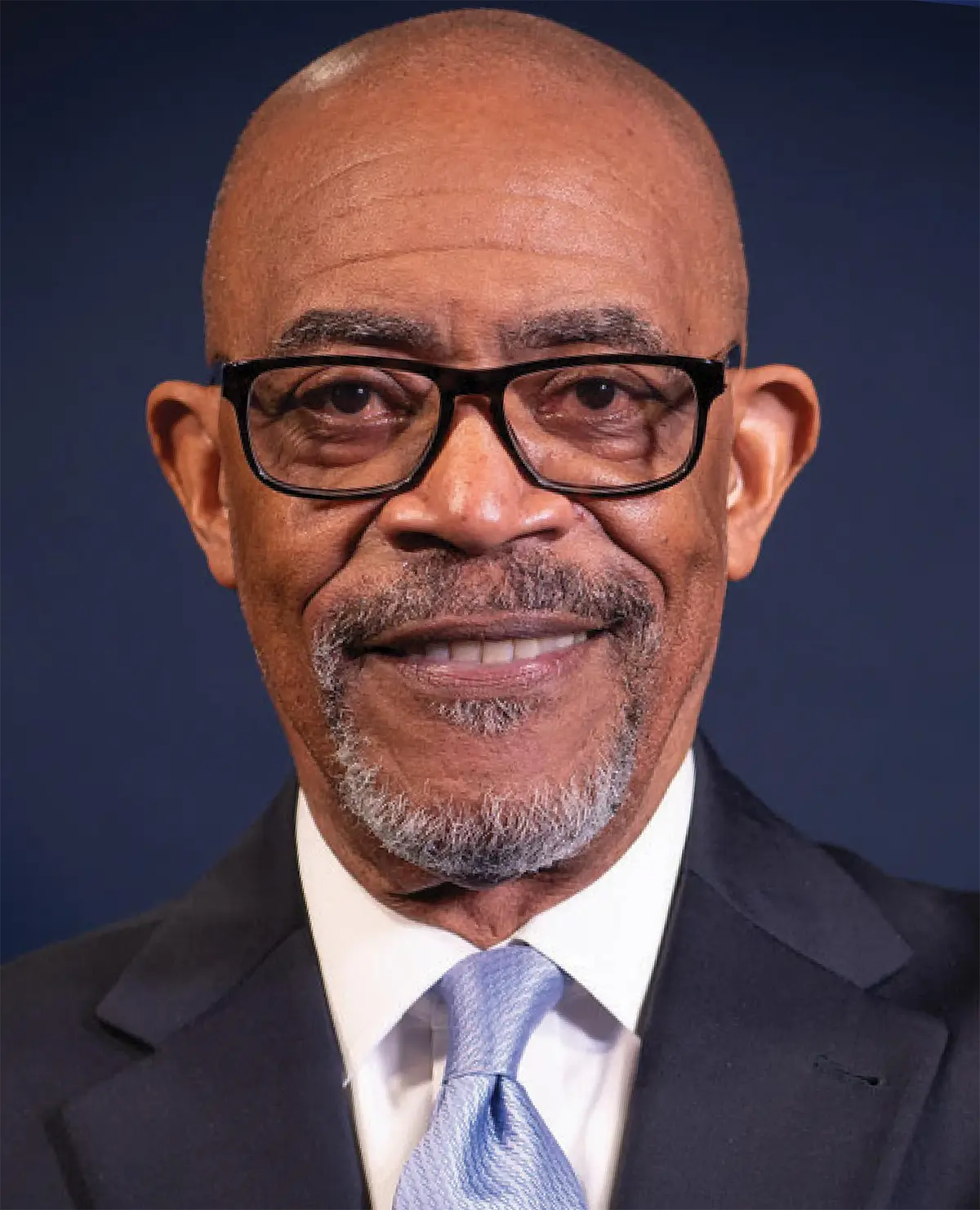 Portrait orientation photograph close-up headshot of Dr. Lester W. Young, Jr. smiling as he is wearing a thin black dark outer frame and dark black dim inner tinted prescription eyeglasses plus has shades of black/grey/white goatee facial beard hair; He is dressed in a dark navy blue business blazer suit, white button-up dress shirt underneath, and light sky blue tie equipped