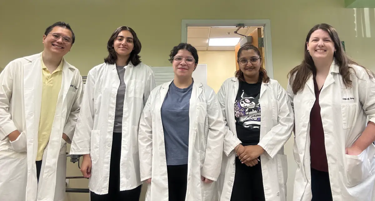 Landscape orientation photograph close-up view of Bor-Shuen Wang, Ph.D., assistant professor; Anastajia Melika ’26; Kaela Collazo ’25; Mandy Bhagwandeen ’25; and Pamela Lovejoy, Ph.D., assistant professor all smiling together standing for a group picture together in their white scientific laboratory coats as they are situated inside a building room of some sort