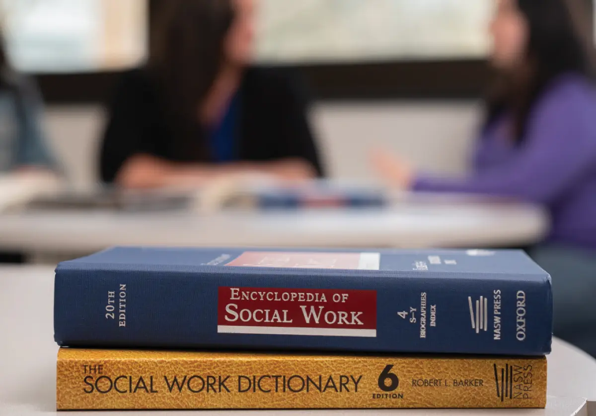 Portrait orientation photograph close-up view of two textbooks stacked on top of each other on a table that are titled as the following respectively: Encyclopedia of Social Work 20th Edition; The Social Work Dictionary Edition 6; Furthermore in the blurred out background behind these textbooks are three women sitting at a different table all talking amongst each other
