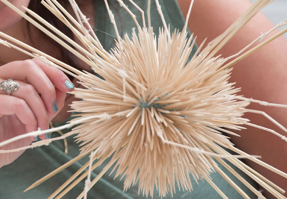 Close-up of a spherical arrangement of wooden skewers resembling a sea urchin, with a person's hand holding one skewer.