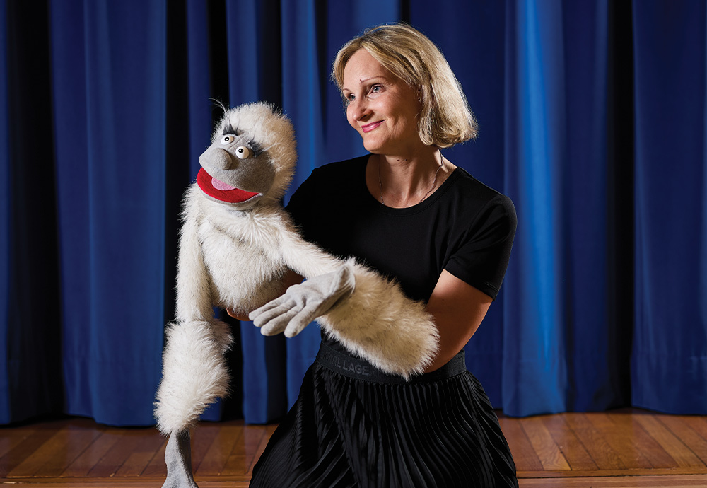 Veronica Kaninska dressed in all black stands on a blue curtained stage wearing an arm puppet