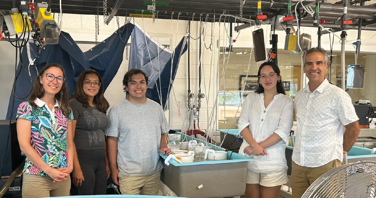 Landscape orientation cropped indoor photograph view of Kestrel Perez, Ph.D., SJNY students Milana Haripersaud ’26, Angel Herrera ’25 and Kleopatra Garo ’26, and Konstantine Rountos, Ph.D. all smiling for a group picture in casual attire standing inside a scientific biology lab space area as there appears to be a whole various assorted equipment around them