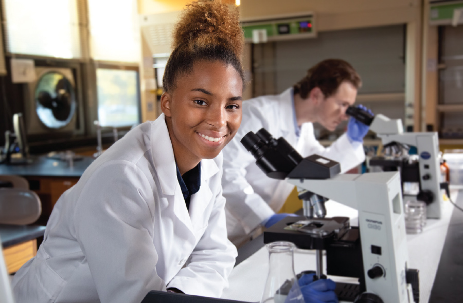 SJNY student smiling in lab at table with microscopes