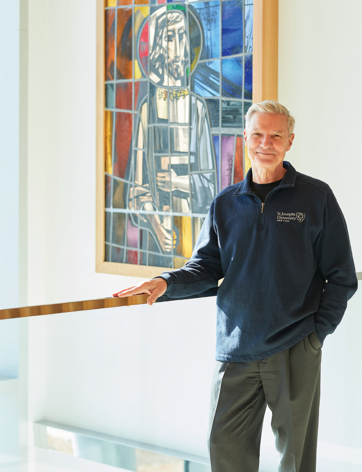 Donald R. Boomgaarden in St. Joseph's Univeristy quarter zip sweater while standing in front of stained glass inspired artwork