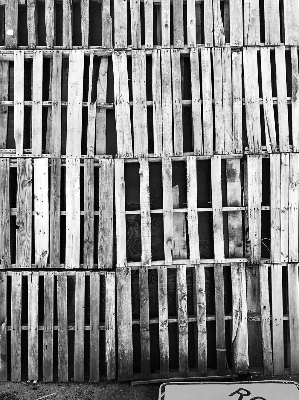 Black and white image of stacked wooden pallets in a grid pattern. A partial sign is visible at the bottom.