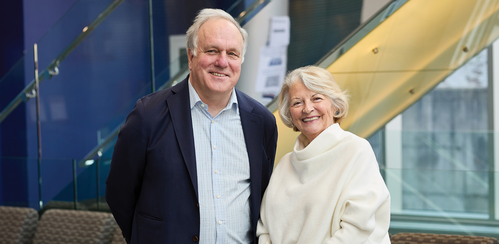 An older man and woman smiling indoors near a glass staircase.