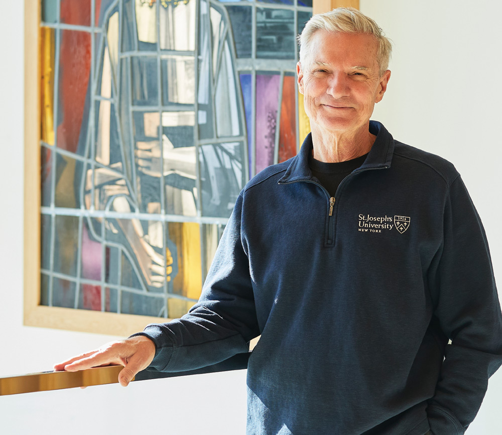 Man in a dark blue pullover standing by a railing with a colorful stained glass window behind him.