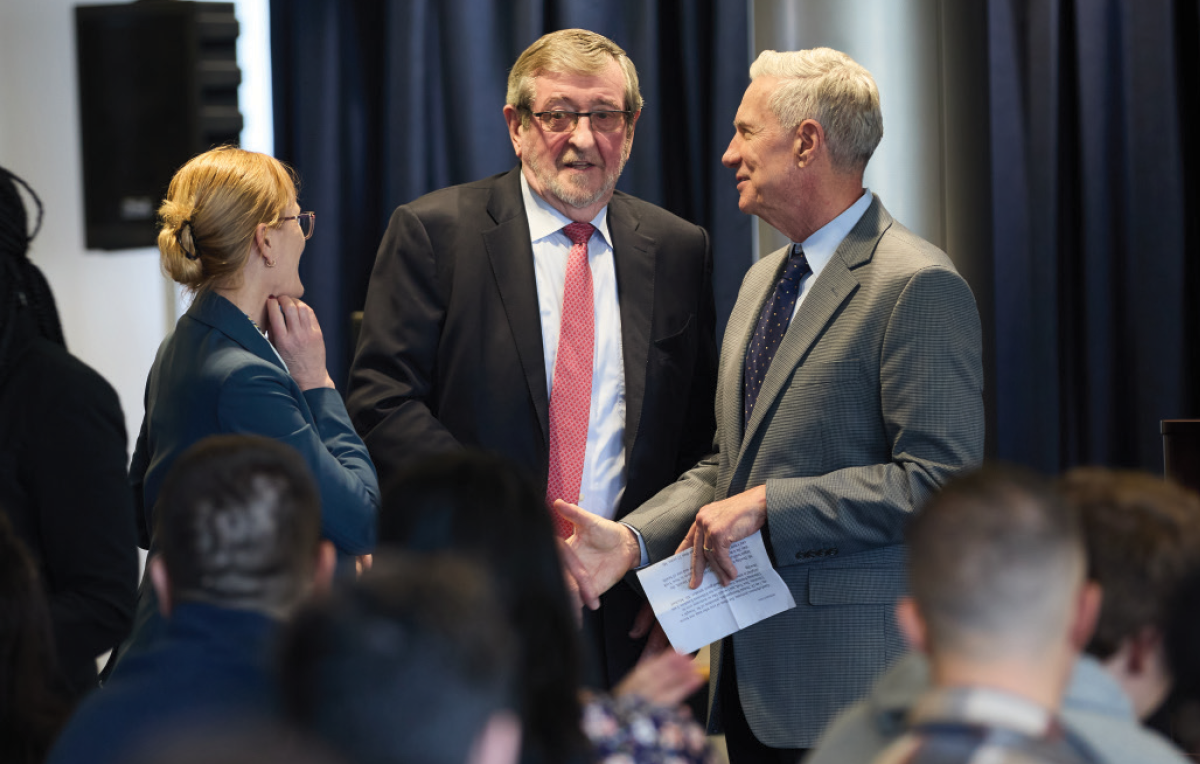 A candid shot of three people engaged in conversation in a professional setting; A man in a dark suit and red tie stands in the center, flanked by a woman in a blue blazer on the left and a man in a gray suit on the right; One of the men pictured here is Michael Dowling of Northwell Health