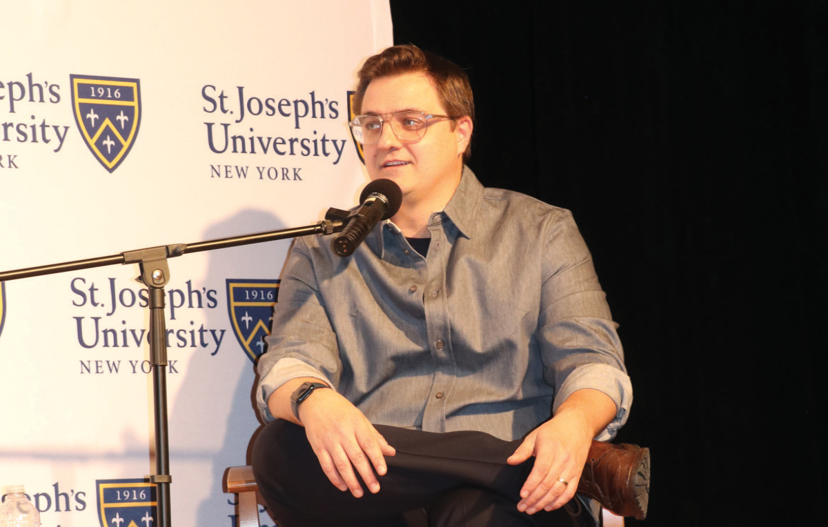 Chris Hayes (from MS NOW), a man in a denim button-up shirt and prescription eyeglasses sits in a chair and speaks into a microphone on a stage; The background features repeated logos for St. Joseph’s University New York