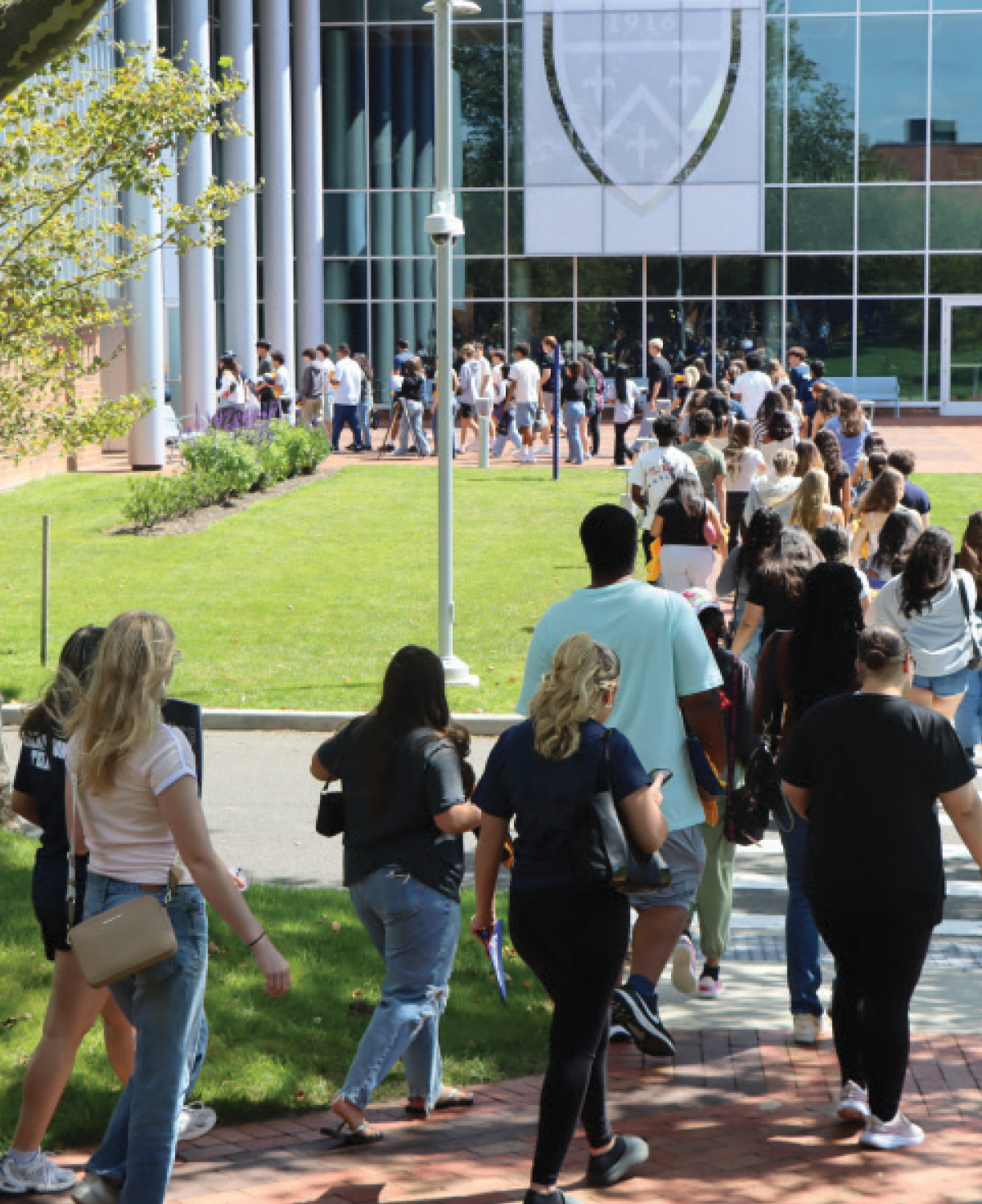 A high-angle shot looking down a long line of students walking toward a large glass St. Joseph's University, New York university building; A large white shield logo with the year 1916 is visible on the glass facade