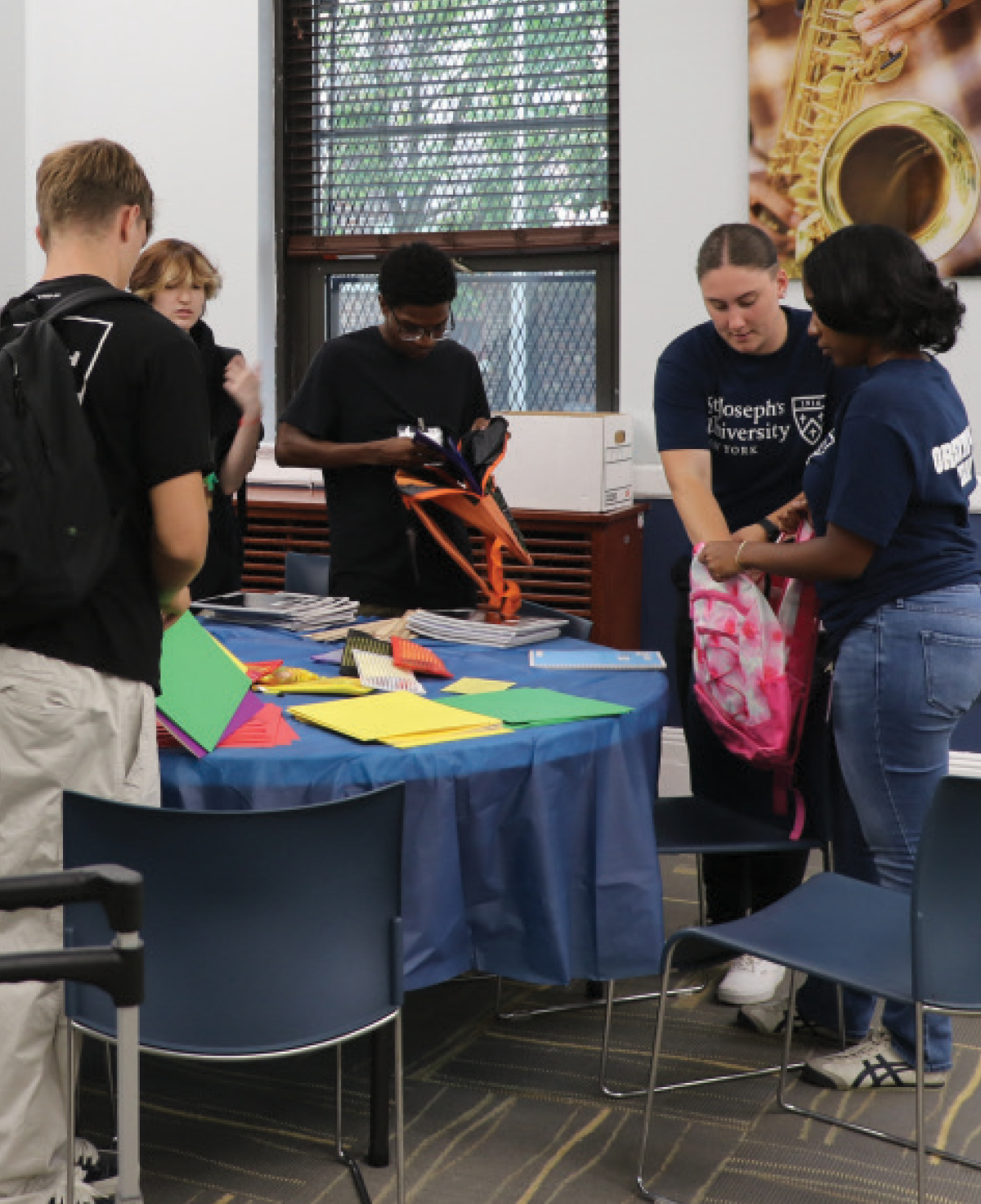 Four young people are gathered standing around a blue circular table in a classroom; They are busy organizing school supplies, including notebooks, folders, and pink backpacks