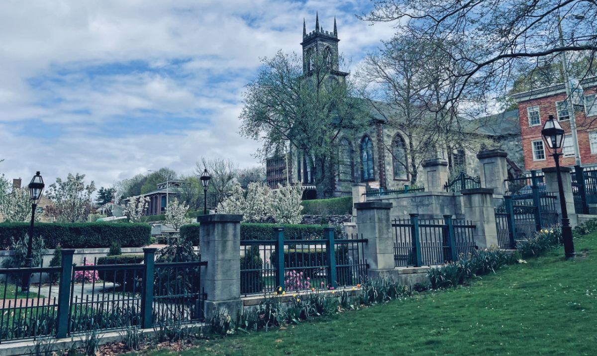 A scenic outdoor view of a stone church building with a tall steeple, partially framed by blooming white trees; A black iron fence runs along a stone wall in the foreground, bordering a green lawn