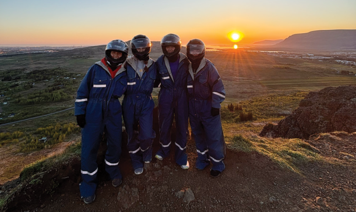 Four people wearing dark blue jumpsuits and matching full-face helmets stand together on a hilltop at sunset; The sun is low on the horizon behind them, casting long shadows across a vast landscape