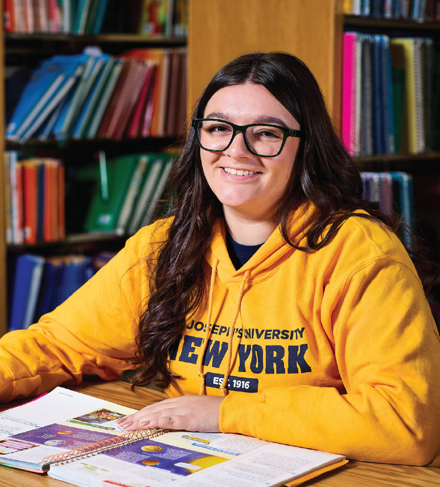 Emma Sasso wearing a yellow SJNY hoodie while sitting with an open book in the school library