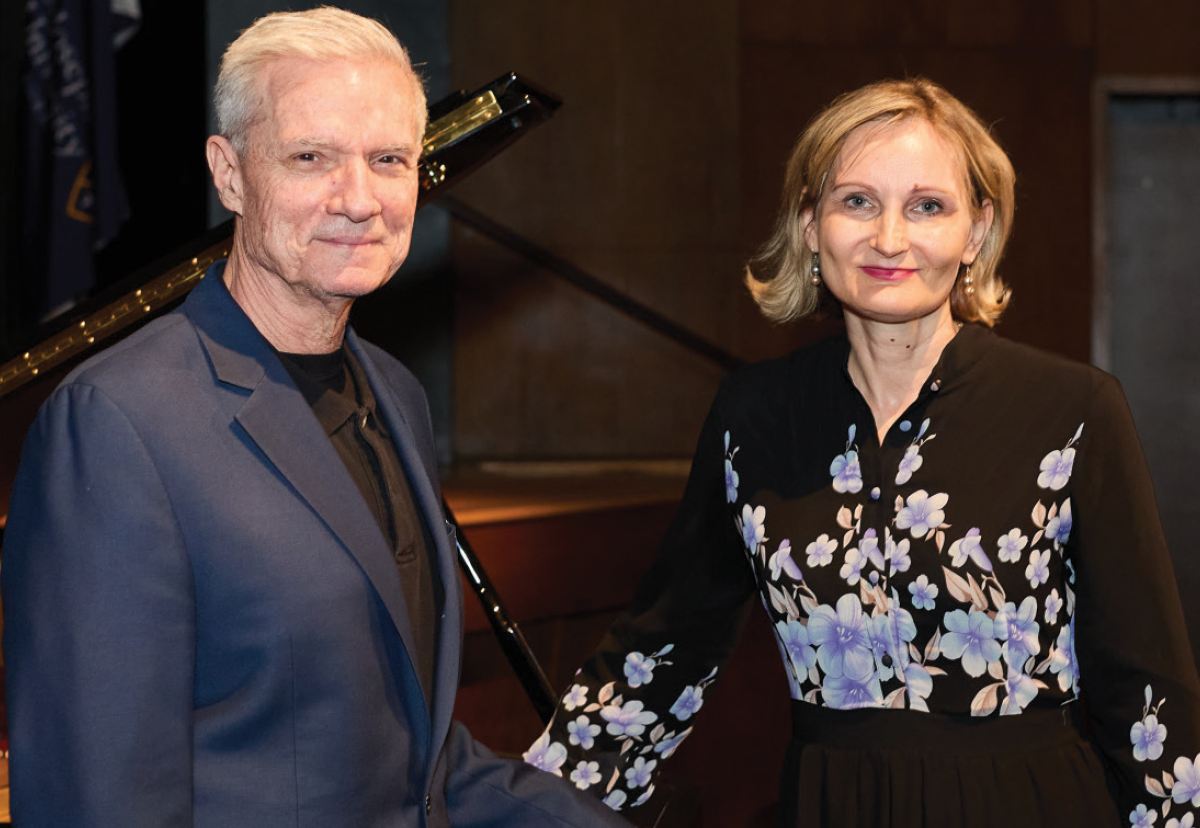 President Donald R. Boomgaarden, Ph.D., a smiling man in a dark navy blue blazer suit and Veronica Kaninska, M.S.Ed, C.T.R.S., a smiling woman in a black floral blouse stand together in front of a grand piano on a stage