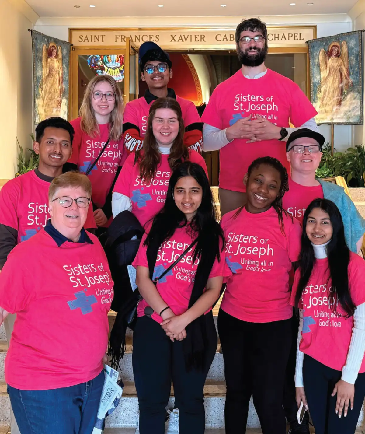 SJNY students posing together during a community service event, wearing bright pink shirts that read 'Sisters of St. Joseph: Uniting all in God's love' inside a historic chapel.