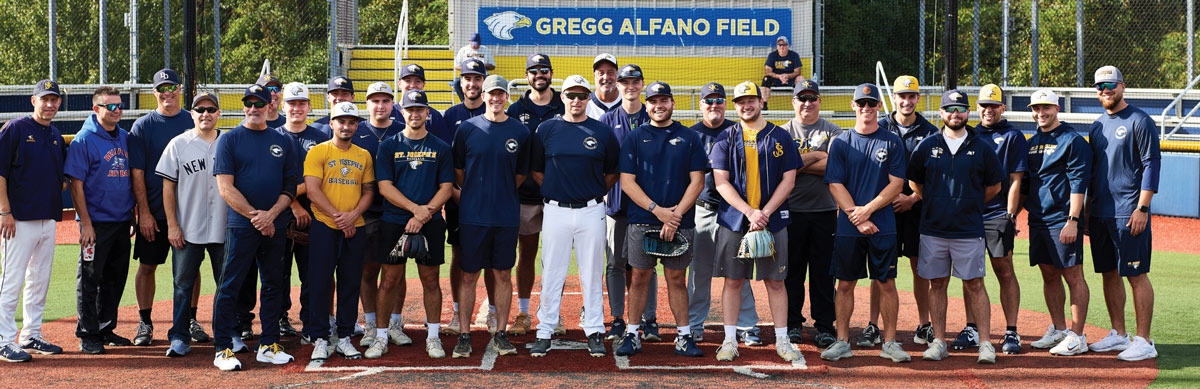 A large group of male baseball players and alumni in St. Joseph’s University navy and gold gear pose together on Gregg Alfano Field for a commemorative team photo.