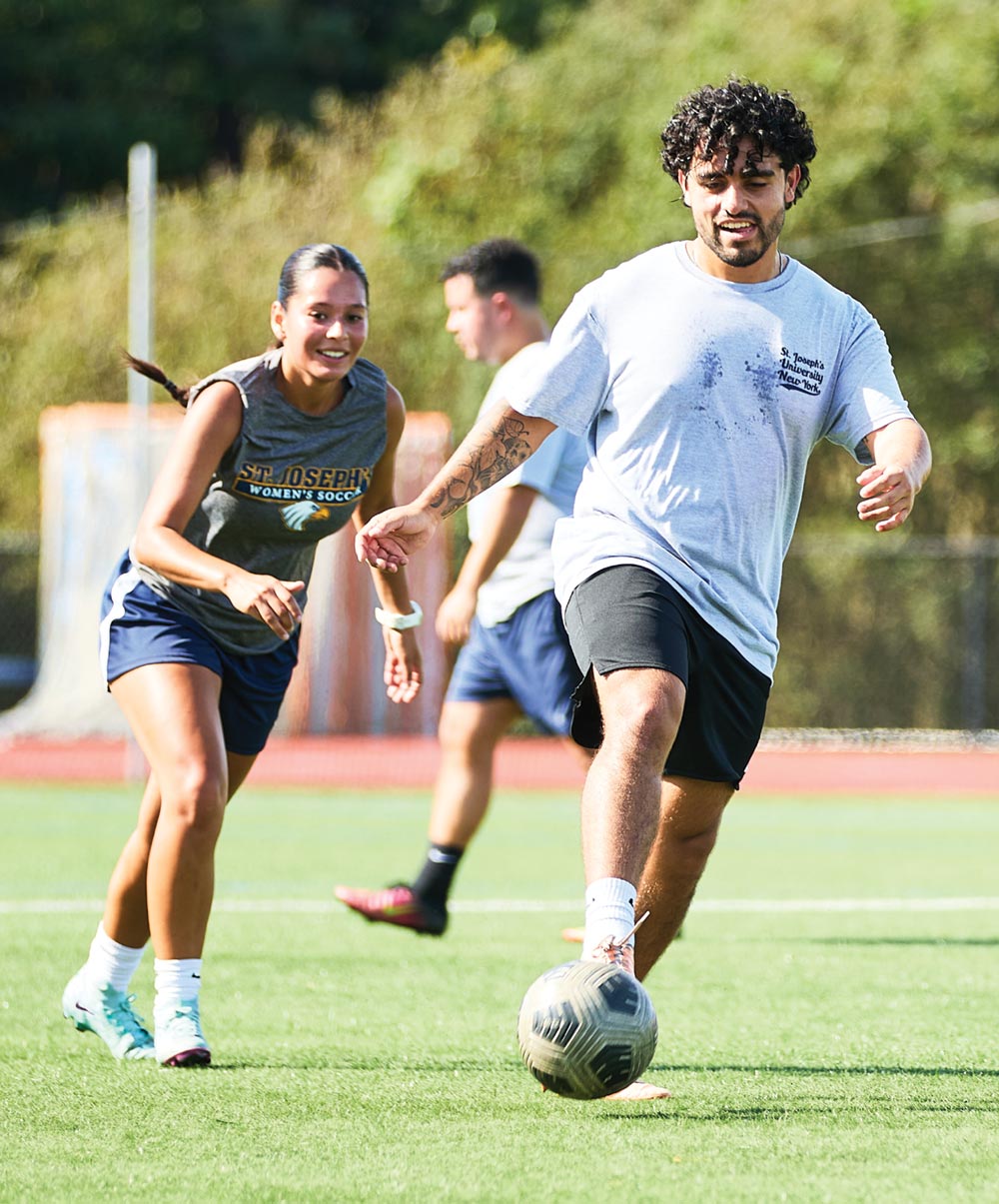 An action shot of an alumni soccer game where a man in a grey St. Joseph’s t-shirt dribbles a soccer ball while a woman in a Golden Eagles jersey defends him on the turf.