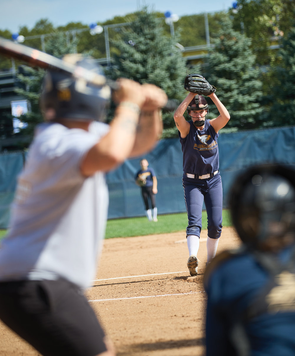 A softball pitcher in a navy Golden Eagles uniform prepares her delivery on the mound, viewed from behind a batter during an alumni game at an outdoor sports complex.