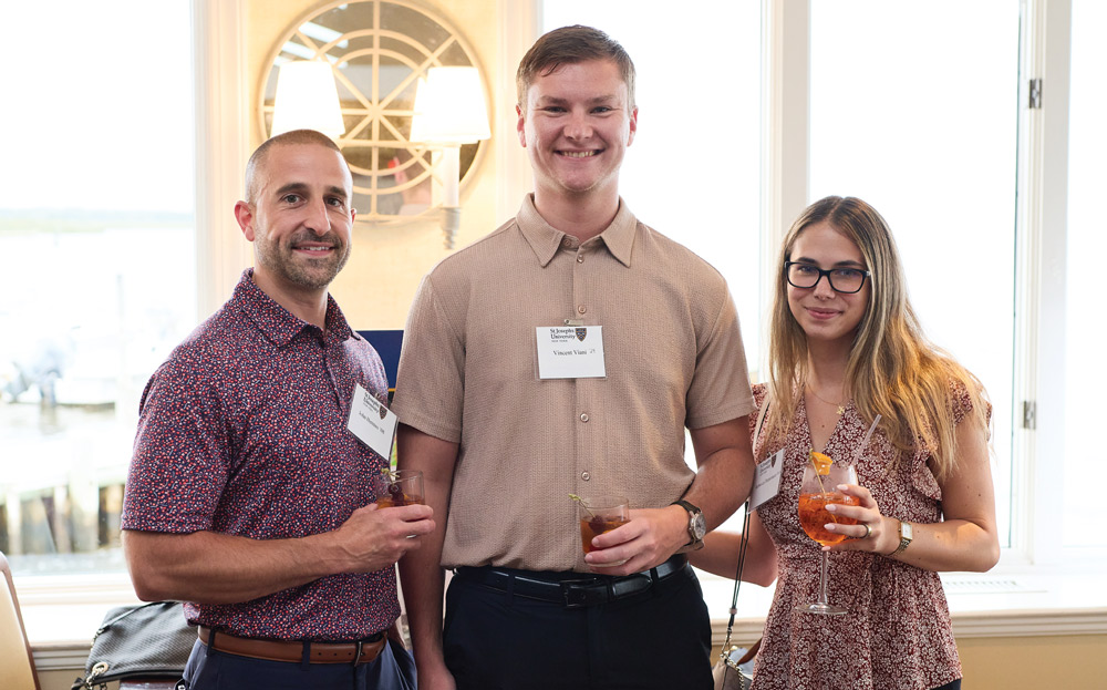 Three alumni, two men and one woman, stand together indoors holding cocktails and wearing name tags while smiling for a photo during a formal University networking event.