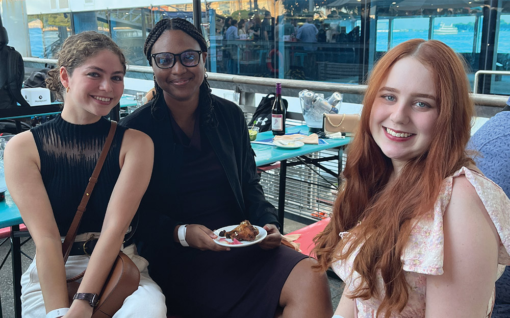 Three young women smile for a portrait while seated at a blue table on an outdoor boat deck, enjoying food and drinks with a scenic waterfront and city skyline behind them.