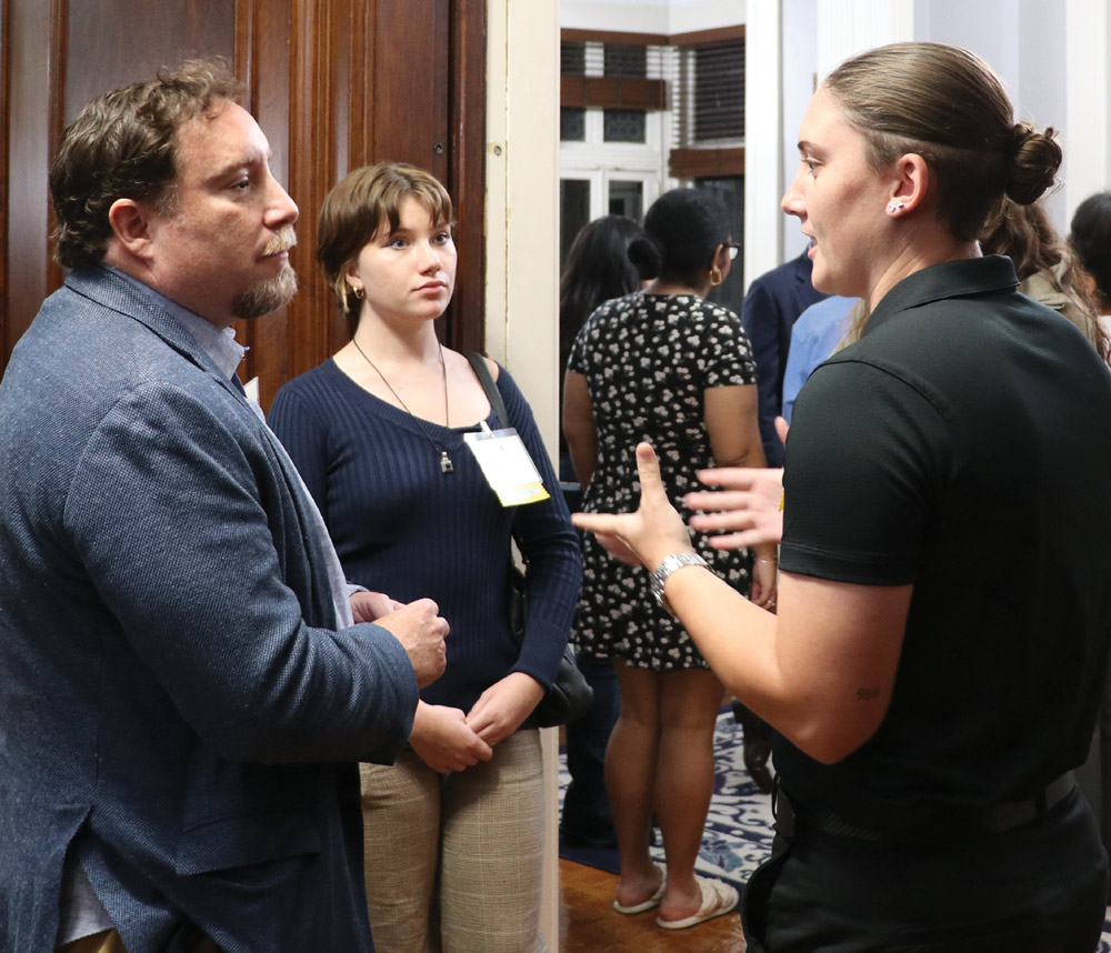 A woman in a black polo shirt gestures while speaking to a man in a blue blazer and a young woman during a crowded indoor alumni networking reception.