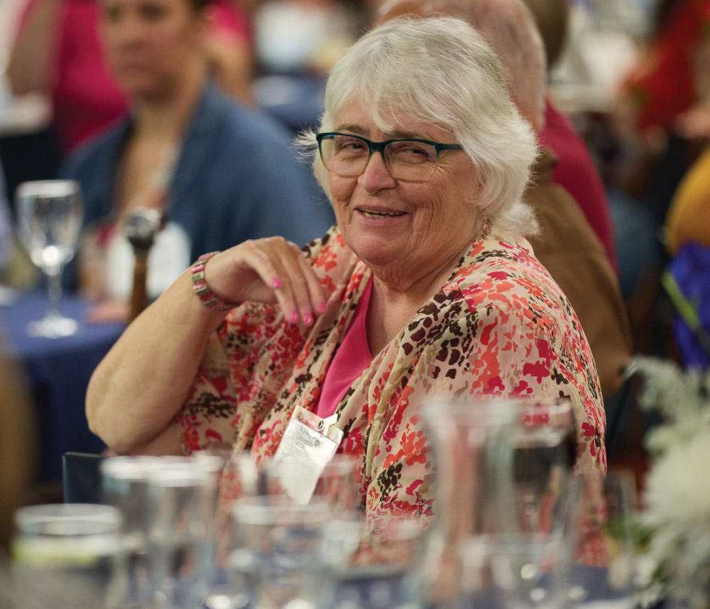 A close-up portrait of a smiling senior alumna with white hair and glasses, wearing a floral print blouse and a name tag while seated at a banquet table during an event.