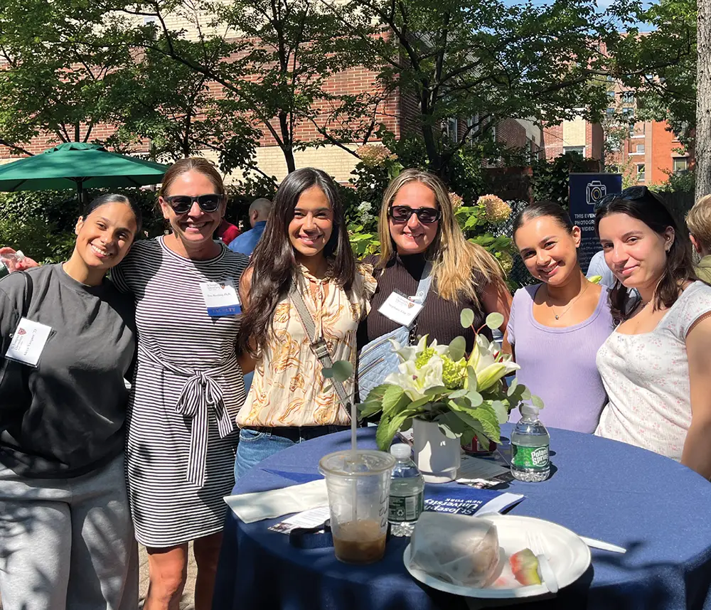 A group of six women, including faculty and alumni, smile together around a high-top table during a sunny outdoor garden reception featuring floral centerpieces.