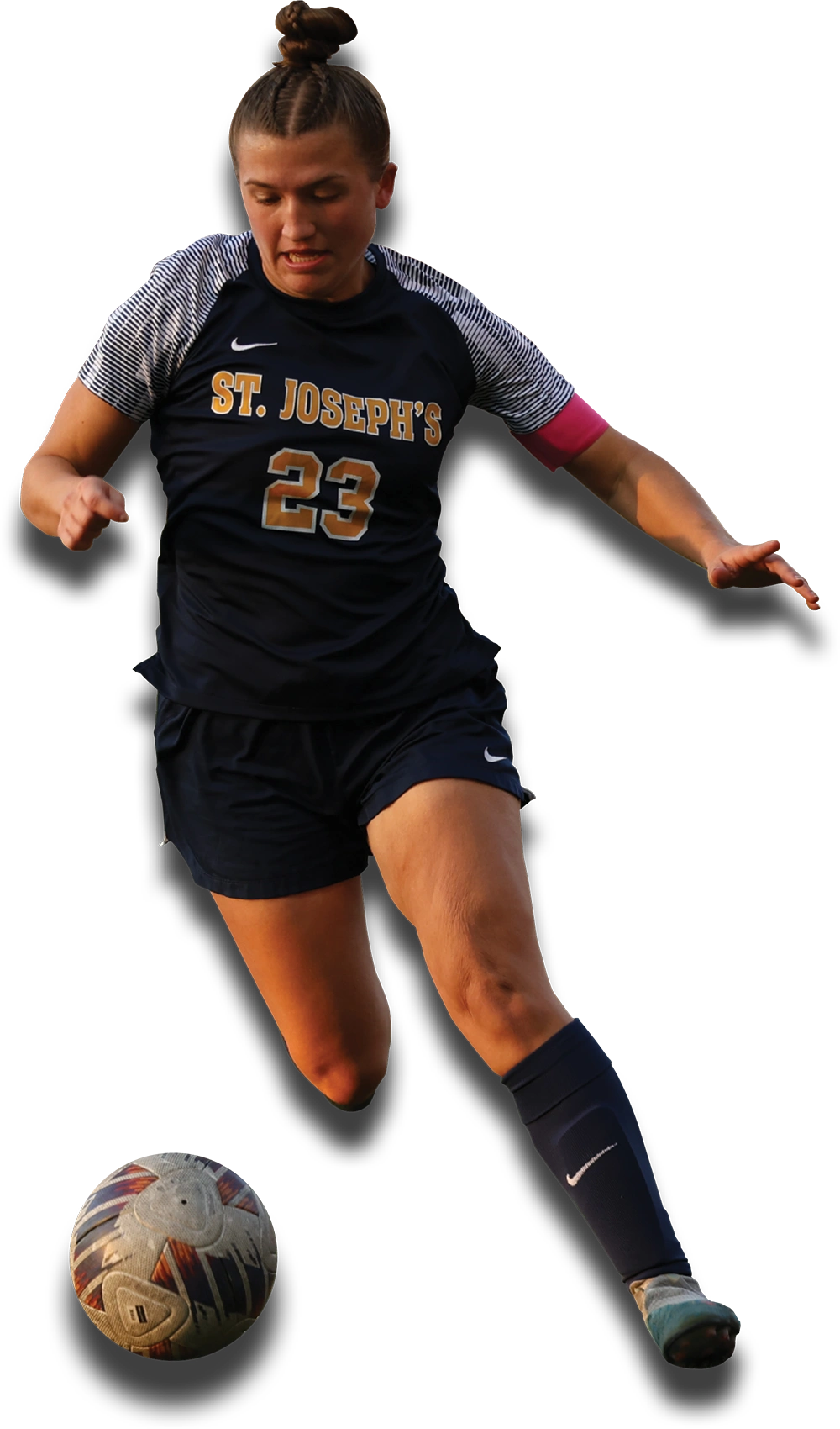 A female St. Joseph’s University soccer player in a dark blue uniform with jersey number 23 dribbles a soccer ball. She has her hair in a bun and wears a pink captain's armband.