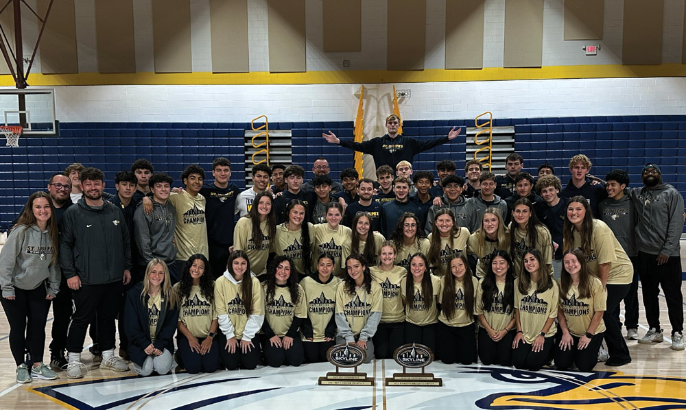 A large group of men’s and women’s soccer players and coaches pose in a gymnasium. Many wear "Skyline Conference Champions" shirts, with two championship trophies placed in the front.