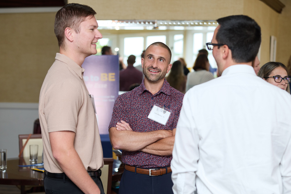 Three men wearing St. Joseph's University name tags engage in a friendly conversation at a reception. One man in a patterned shirt stands with arms crossed, smiling at his peer.