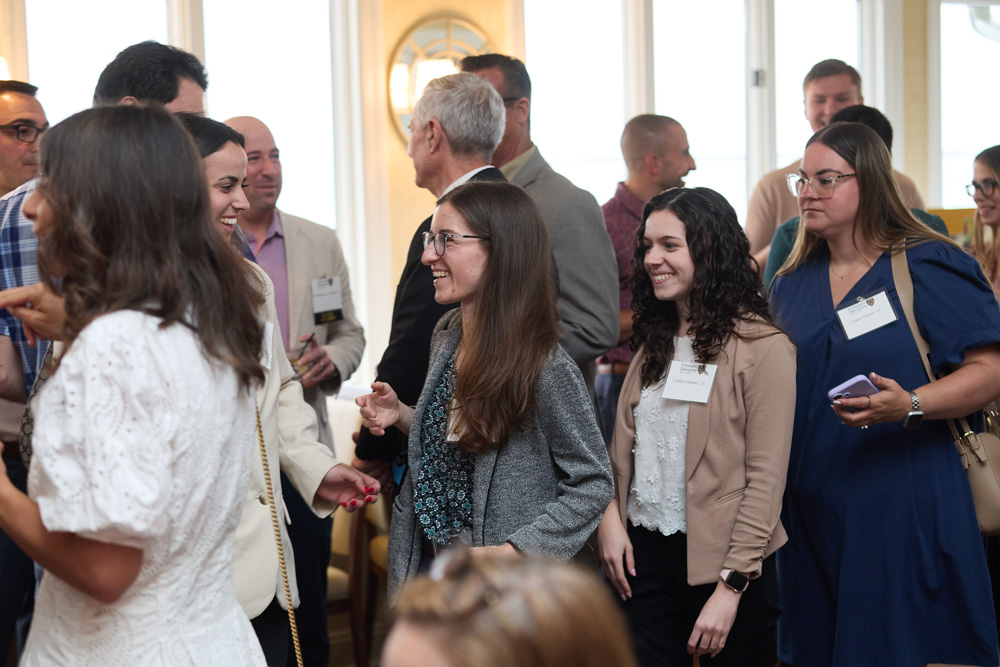 A candid, lively scene of guests networking and laughing at a university alumni event. People are dressed in business casual attire and wearing name tags.