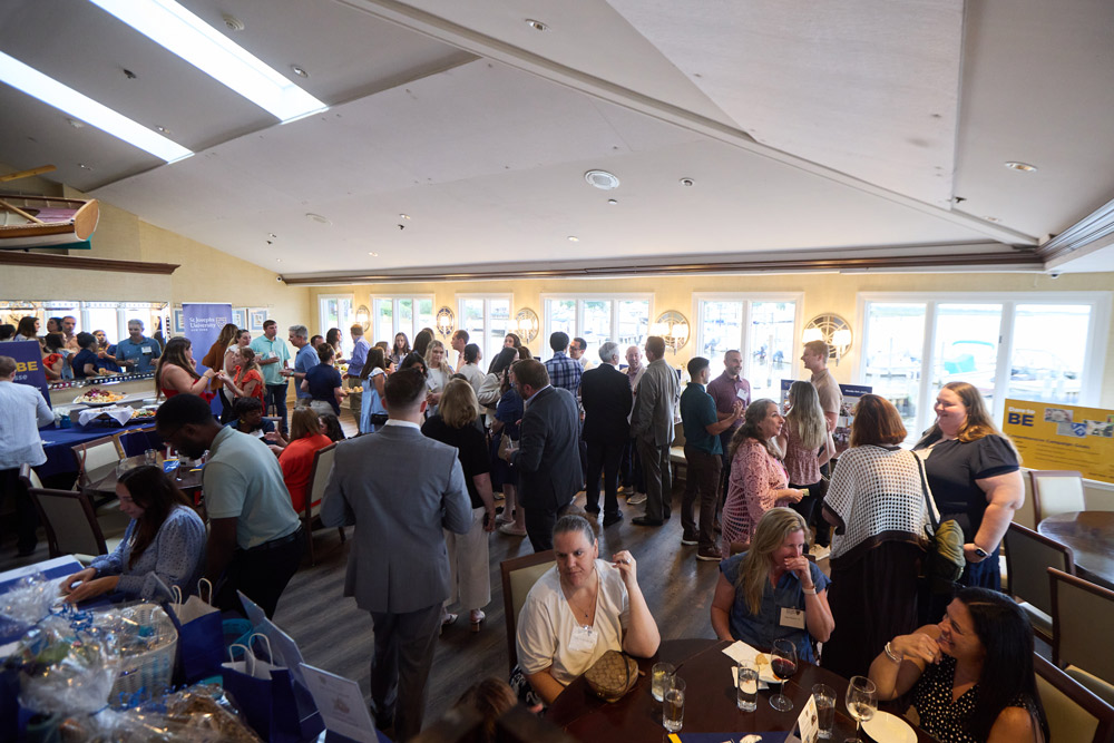 A wide-angle view of a crowded reception room filled with alumni talking. The space features large windows, nautical decor, and tables with blue tablecloths and gift bags.