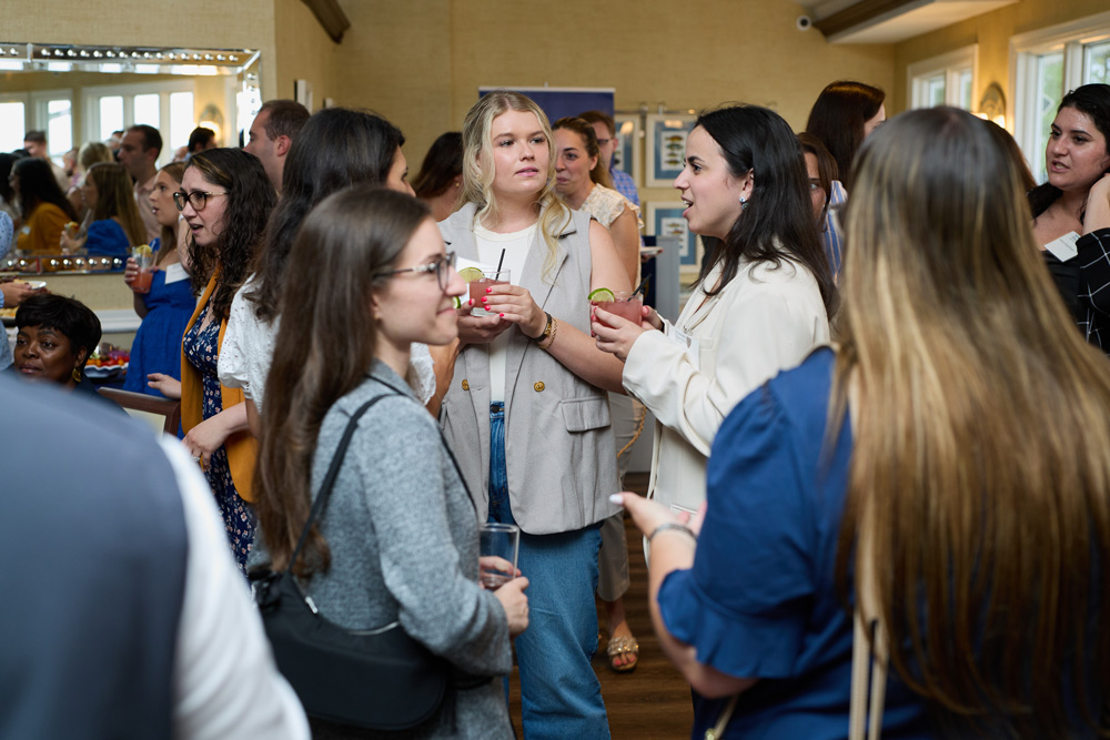 A group of young women engaged in conversation at a social event. They are holding pink cocktails and dressed in stylish, casual professional attire in a sunny room.