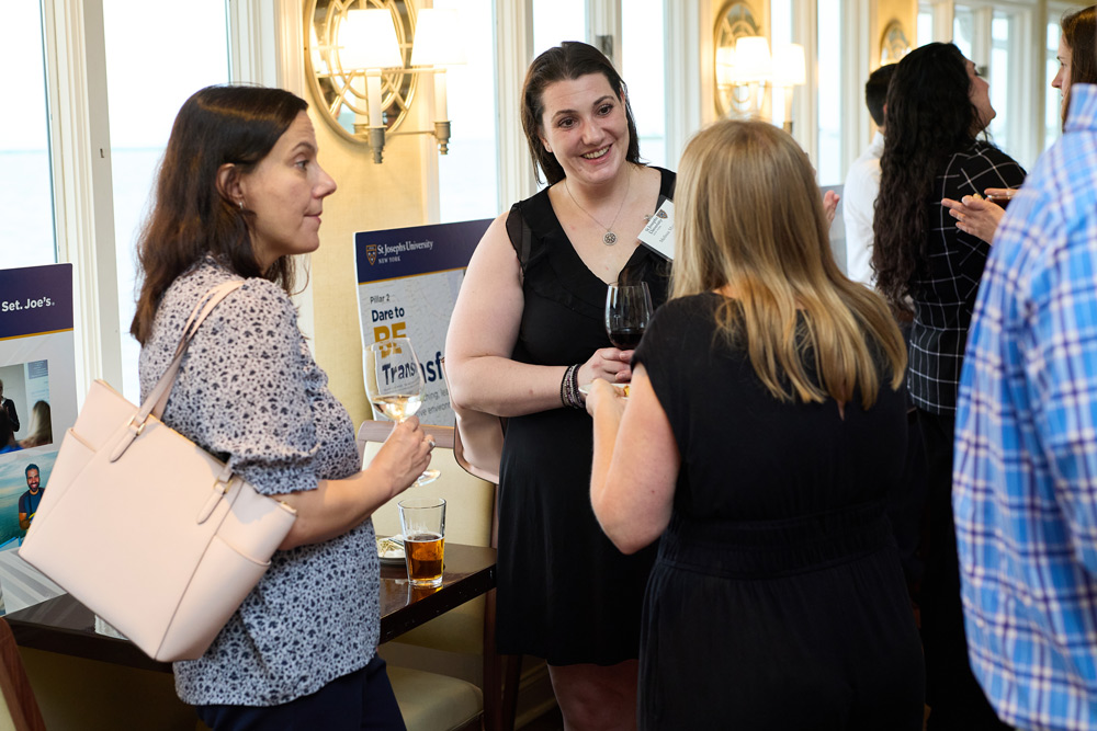 Three women stand in a circle talking and smiling while holding glasses of wine and beer. A "Dare to Be" university campaign poster is visible in the background.