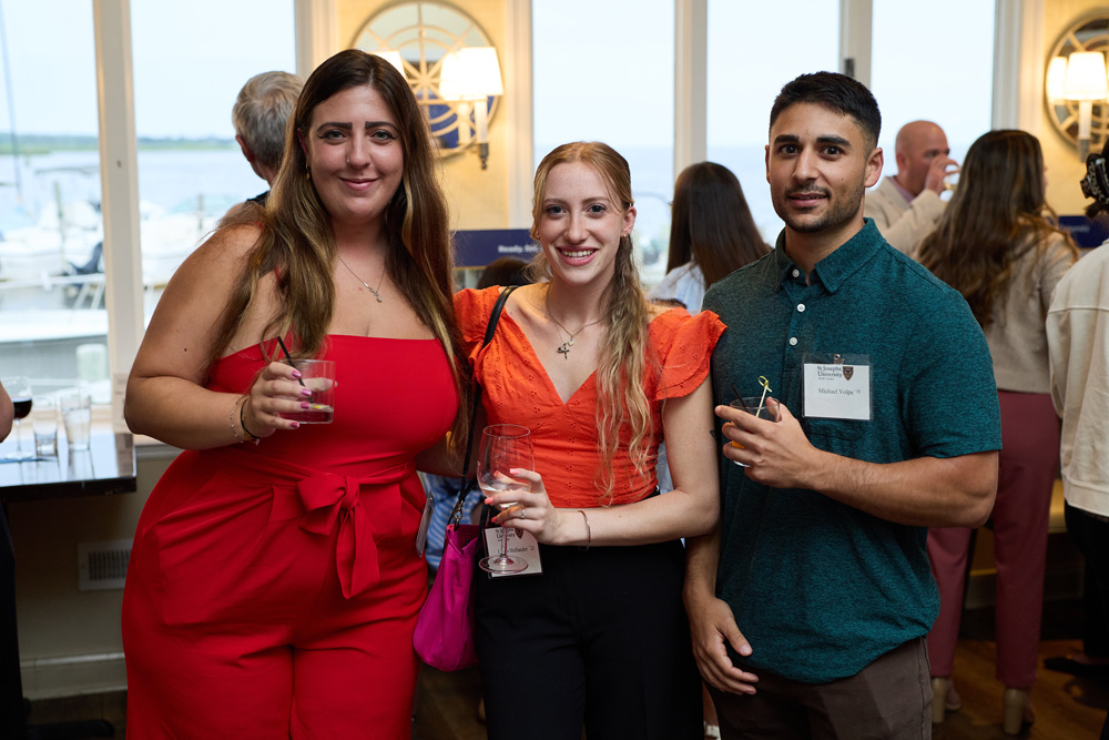 Two women in vibrant red and orange outfits and a man in a teal polo shirt pose for a group photo while holding drinks at a bright waterfront alumni mixer.