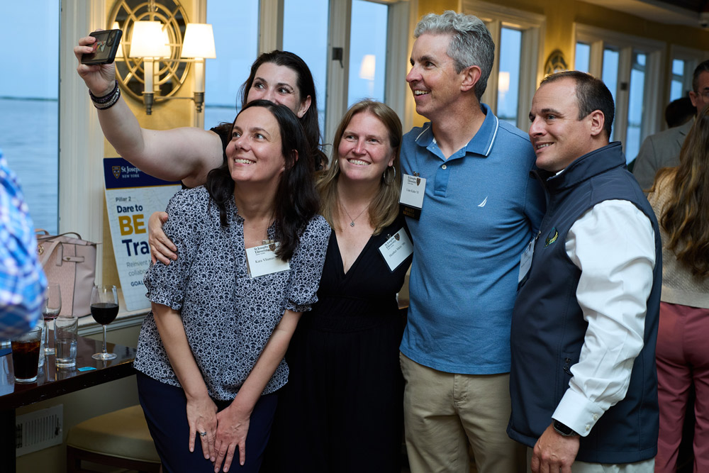 A group of five alumni huddle together and smile for a selfie being taken on a smartphone. They are standing in a well-lit room with a scenic waterfront view in the background.