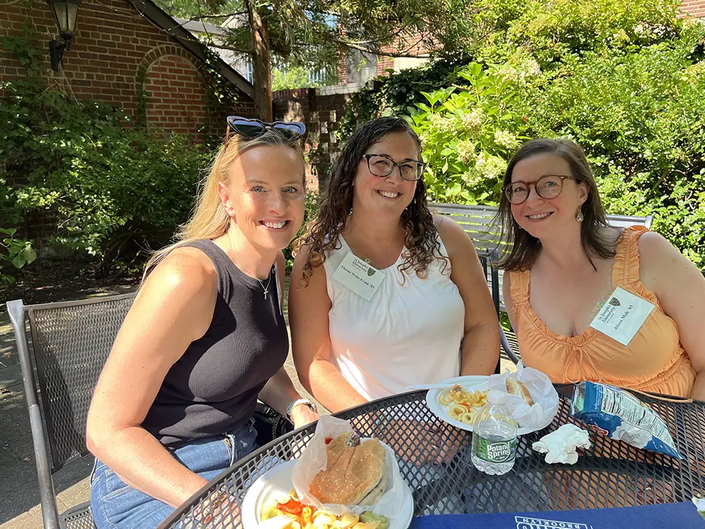 Three women smile for a photo while seated at an outdoor mesh table with lunch. They are wearing St. Joseph's name tags in a sunny, green garden setting.