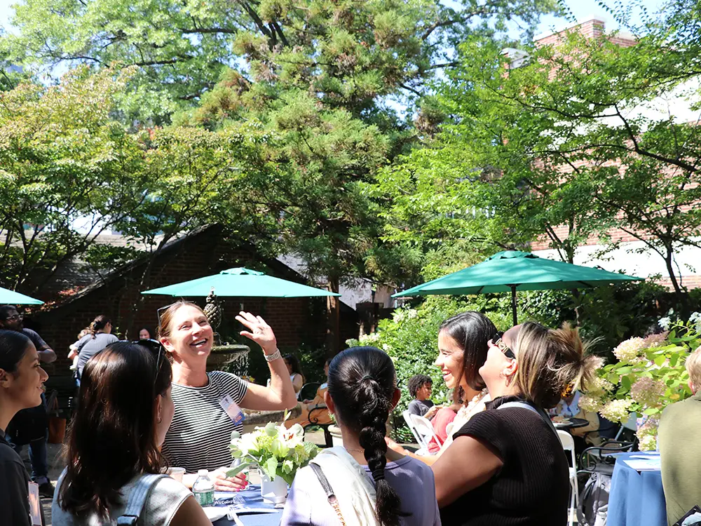 A group of women laugh and talk around a table at an outdoor garden party. Sunlight filters through the trees, creating a bright and joyful atmosphere.
