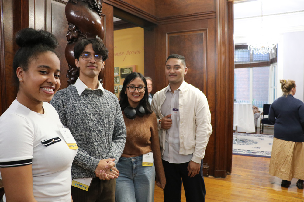 Four students stand together in a wood-paneled room during a networking event. They are dressed in casual professional attire and wearing "Student" name tags.
