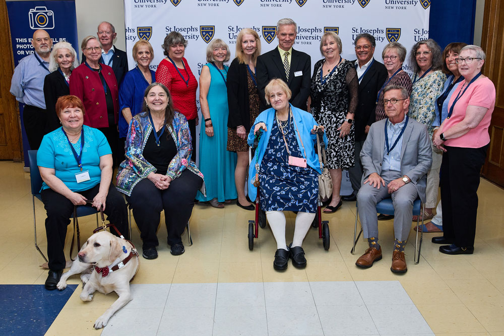 A large group of alumni pose together in front of a St. Joseph's University backdrop. The group includes a woman in a wheelchair and a yellow lab service dog wearing a red bow tie.