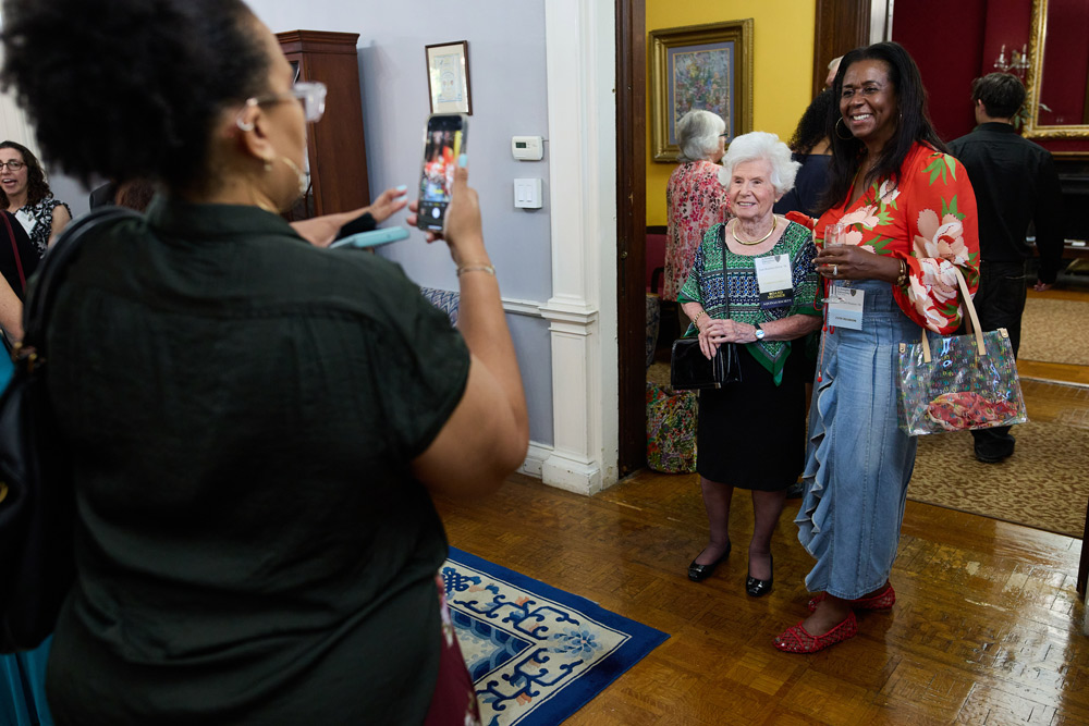 A woman in a floral blouse and denim skirt smiles as a friend takes her photo on a smartphone. They are standing in a classic, well-decorated room during a formal social.