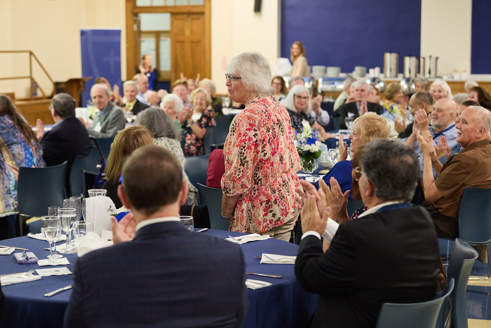 A woman in a floral shirt stands to be recognized at a formal luncheon. Surrounding guests at circular tables with blue linens clap and smile in support.