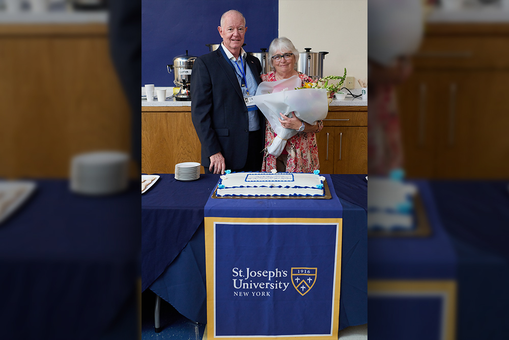 A man in a suit and a woman holding a large bouquet stand behind a blue-clothed table featuring a celebratory sheet cake with the St. Joseph's University New York logo.